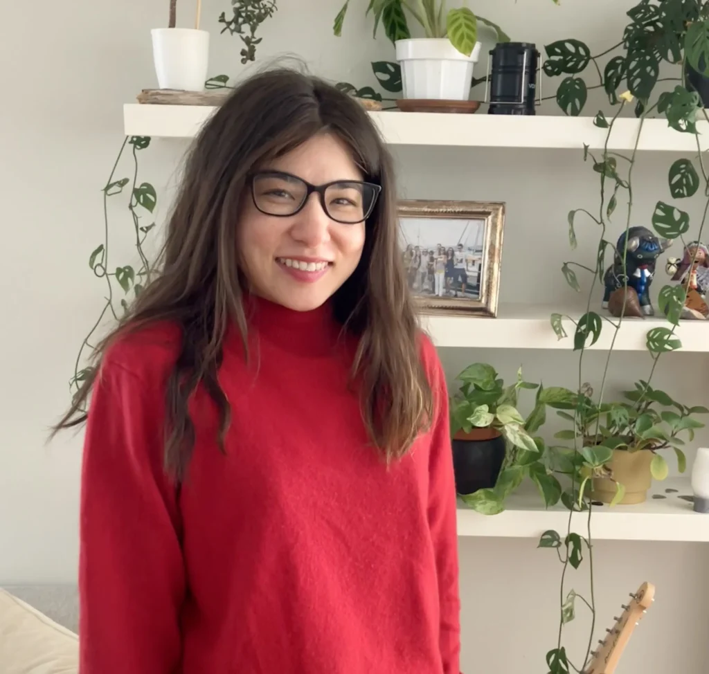 A person in a red sweater stands in front of a shelf adorned with plants, photo frames, and decorative figurines, creating a cozy atmosphere.