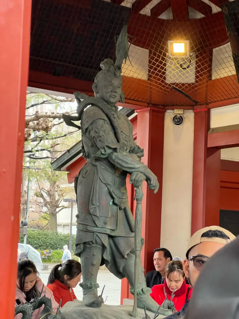 A bronze statue of a warrior stands majestically in a traditional shrine, surrounded by people admiring the intricate details.