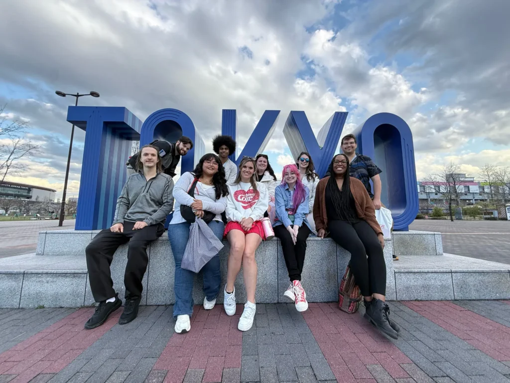 Group of diverse people posing in front of a large, stylized "TOKYO" sign.