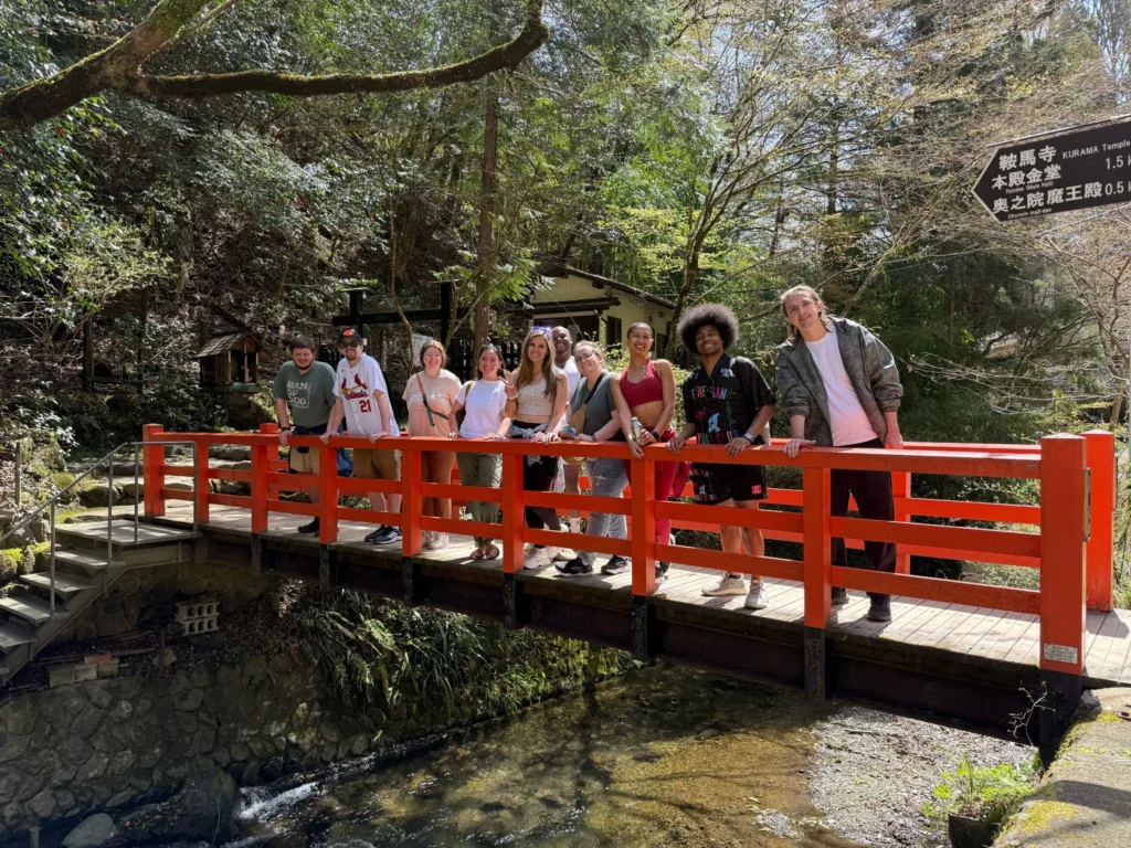 Group of people stands on a vibrant red wooden bridge over a small stream in a lush, green Japanese forest.