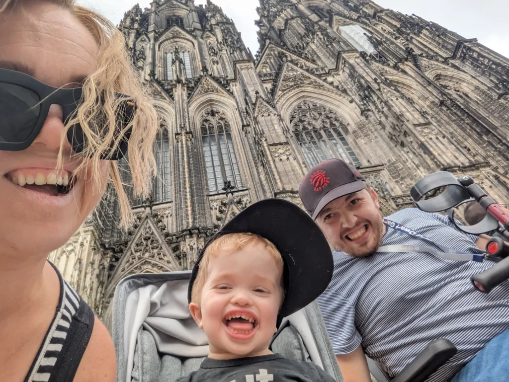A smiling family with a young child in a stroller takes a selfie in front of a large, ornate Gothic cathedral. The child and adults appear happy and are wearing casual clothes. The cathedral towers in the background.