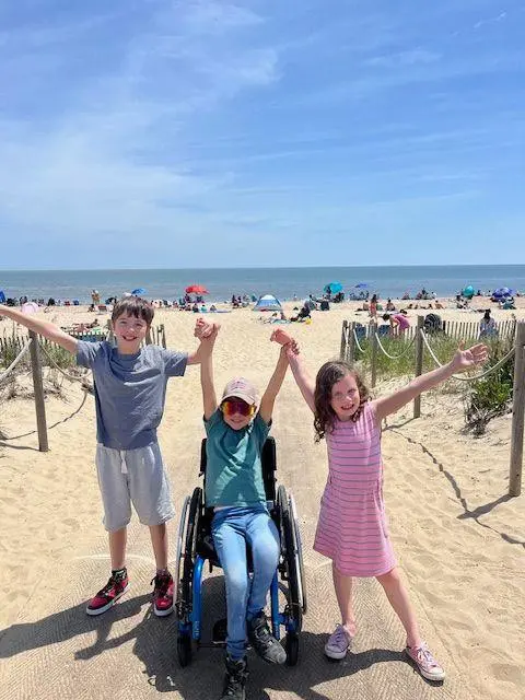 Three children joyfully pose on the beach, one in a wheelchair, with umbrellas and beachgoers in the background under a clear blue sky.