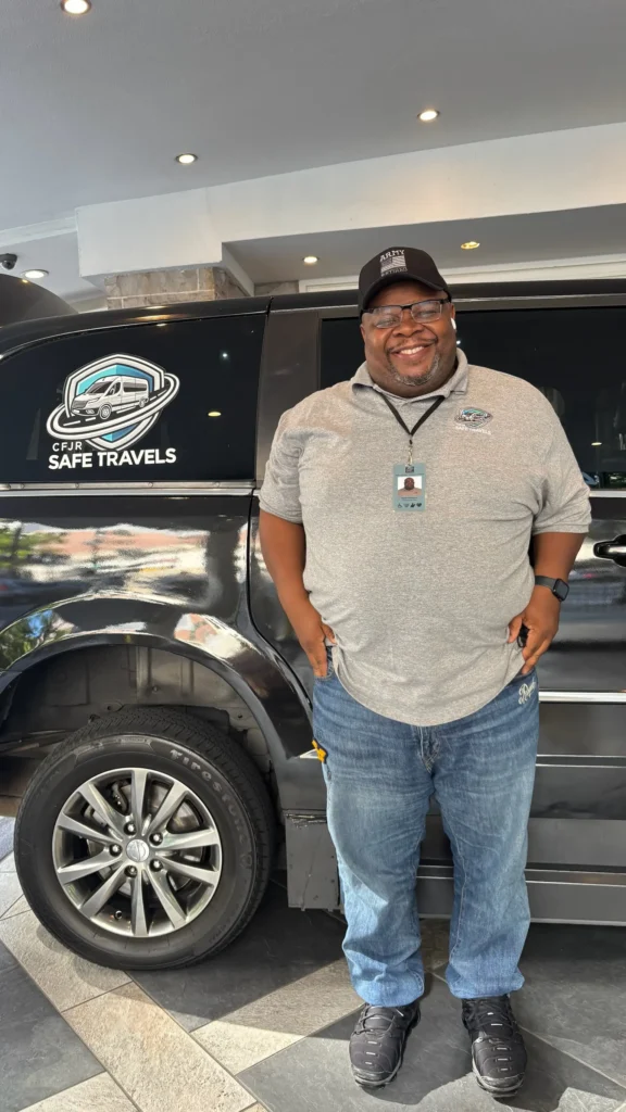 A smiling man standing next to a black vehicle with a logo that reads "Safe Travels." He is wearing a gray polo shirt and a cap, with blue jeans, in a well-lit indoor setting.