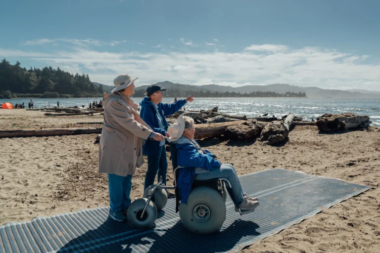 Two women assist a third in a wheelchair on a sandy beach, pointing towards the water under a clear blue sky with distant mountains.