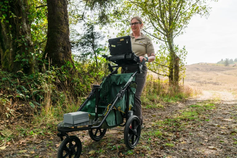 A person stands on a path in a natural setting, pushing a wheeled cart with a laptop and equipment for data collection.