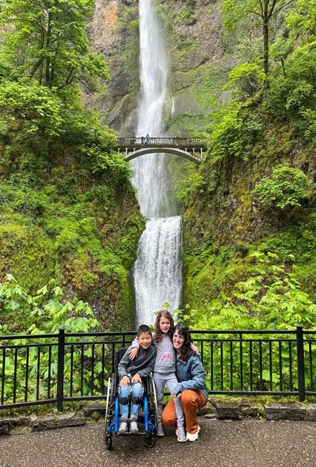 A scenic view of a lush green landscape featuring a tall waterfall and a bridge, with three people posing in the foreground.