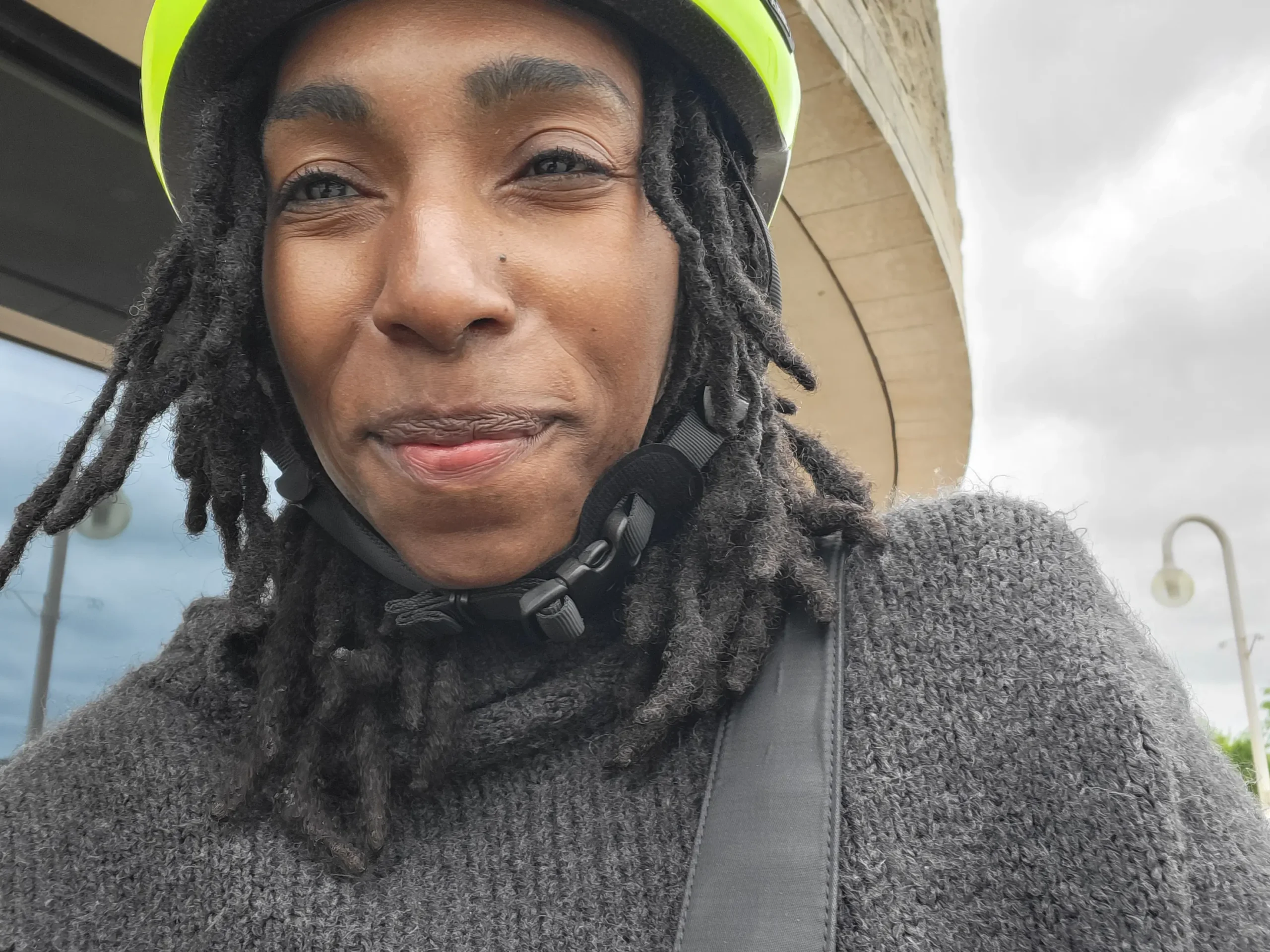 A person wearing a gray sweater and a bright yellow helmet stands outdoors, with a background of cloudy skies and modern architecture.