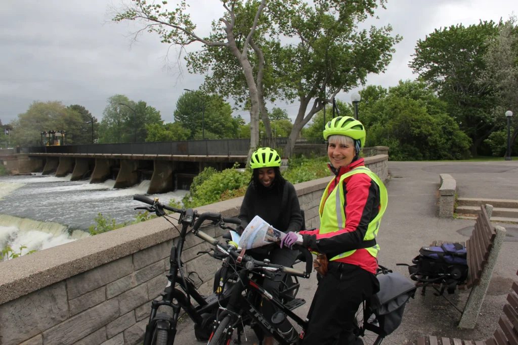 Two cyclists in bright helmets stop by a river, studying a map near a dam and lush greenery under a cloudy sky.