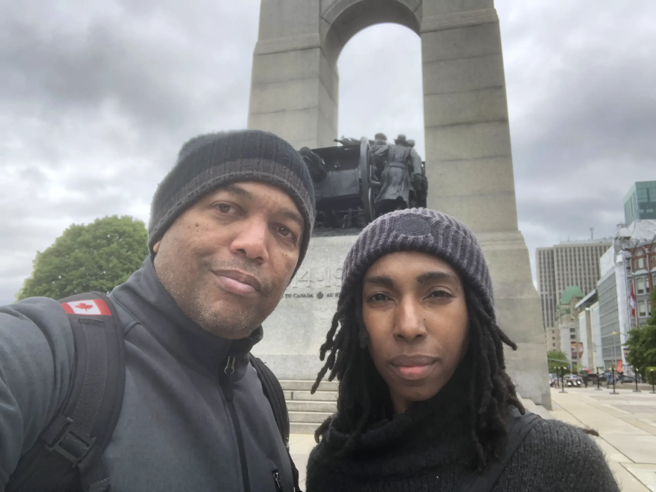 A man and a woman standing in front of a tall war monument under a cloudy sky, showcasing a blend of nature and urban scenery.