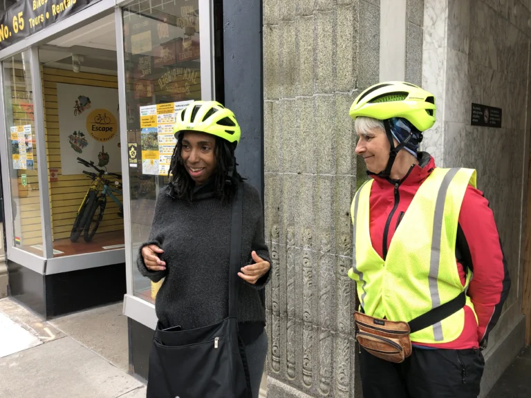 Two cyclists stand outside a bike rental shop, wearing bright yellow helmets and discussing their plans.
