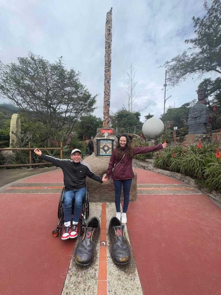 Smiling couple at the equator line monument in Ecuador, with one person in a wheelchair and the other standing, symbolizing inclusive travel and accessibility.