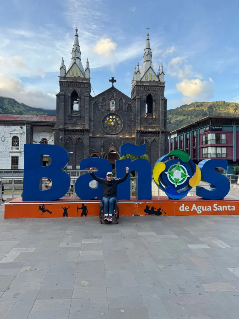 A person in a wheelchair smiles with arms raised in front of large blue Baños de Agua Santa letters, with a historic church and mountains in the background under a partly cloudy sky.