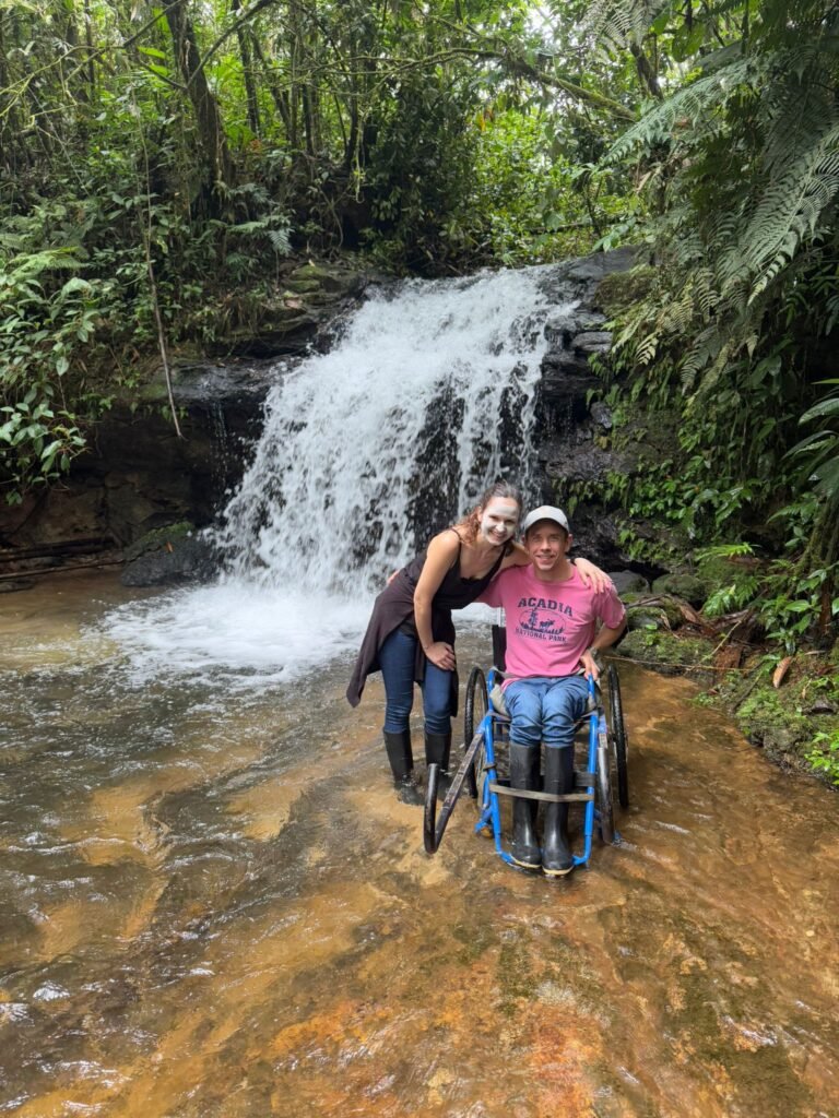 A woman and a man in a wheelchair smile together in front of a small waterfall surrounded by lush green foliage. They are standing in shallow water and both wear rubber boots.