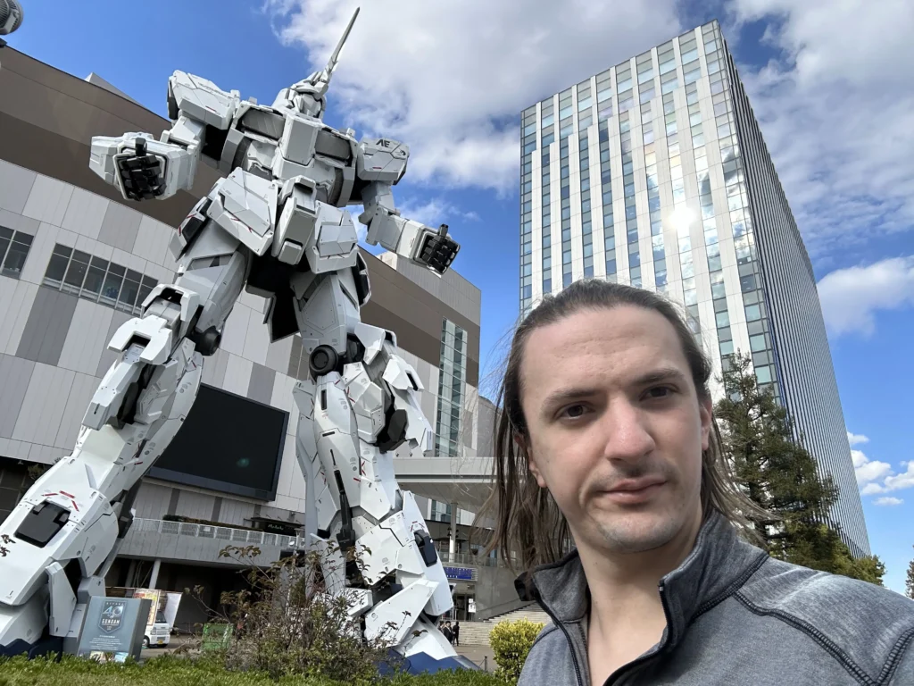 A towering white robot statue stands against a blue sky, next to a modern glass building, with greenery in the foreground.