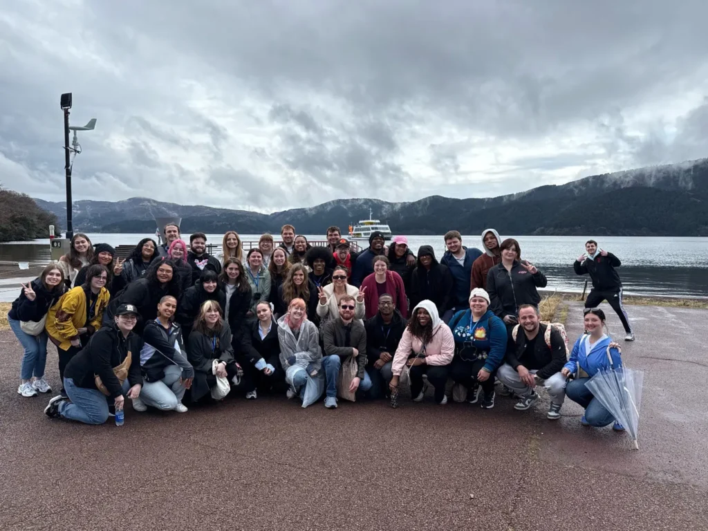 A large group of people gathers outdoors by a lake, with mountains in the background under a cloudy sky.