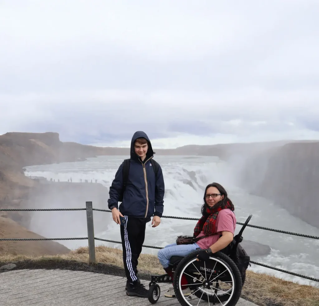A woman in a wheelchair and a standing Boy standing at the edge of a waterfall viewpoint, with mist rising and dramatic cliffs in the background.