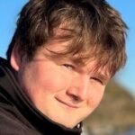 Close-up of a young man smiling outdoors on a sunny day, with blue sky and natural landscape in the background