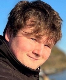 Close-up of a young man smiling outdoors on a sunny day, with blue sky and natural landscape in the background
