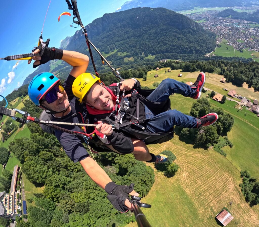 Two people wearing helmets are tandem paragliding high above a green landscape with hills, fields, and scattered buildings. One person holds a selfie stick, capturing their flight and the scenic view below.