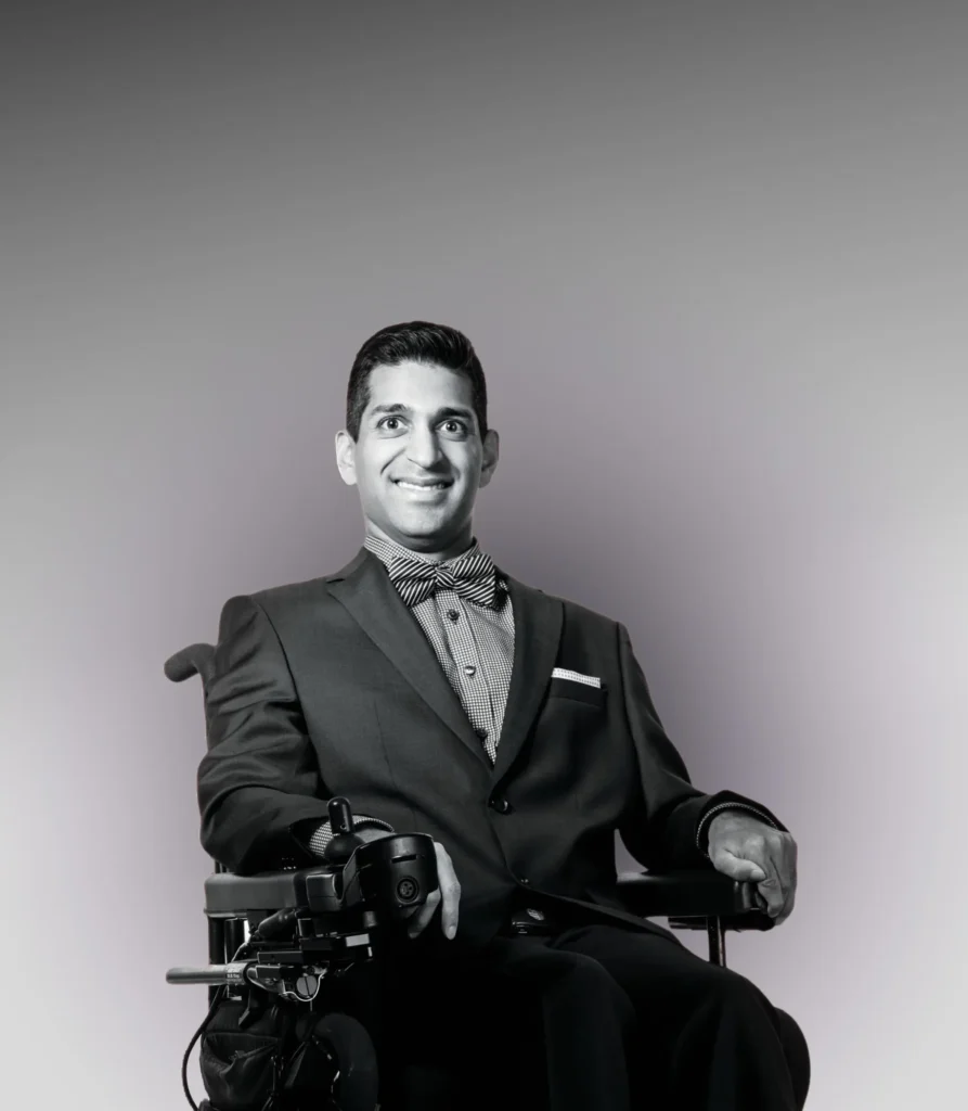 A man in a suit and bow tie sits in a power wheelchair, smiling confidently against a gradient gray background.