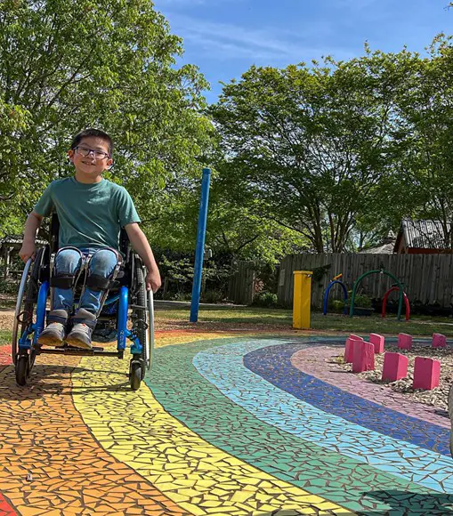 A child in a wheelchair navigates a vibrant, rainbow-colored playground path surrounded by greenery and play structures.