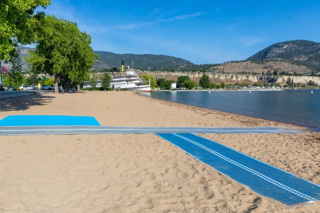 A sandy beach with blue accessible mats leading toward the water, green trees on the left, hills in the background, and a docked paddlewheel boat near the shore under a clear blue sky.
