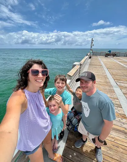 A family group selfie on a sunny pier overlooking the ocean, with blue skies and fluffy clouds in the background.