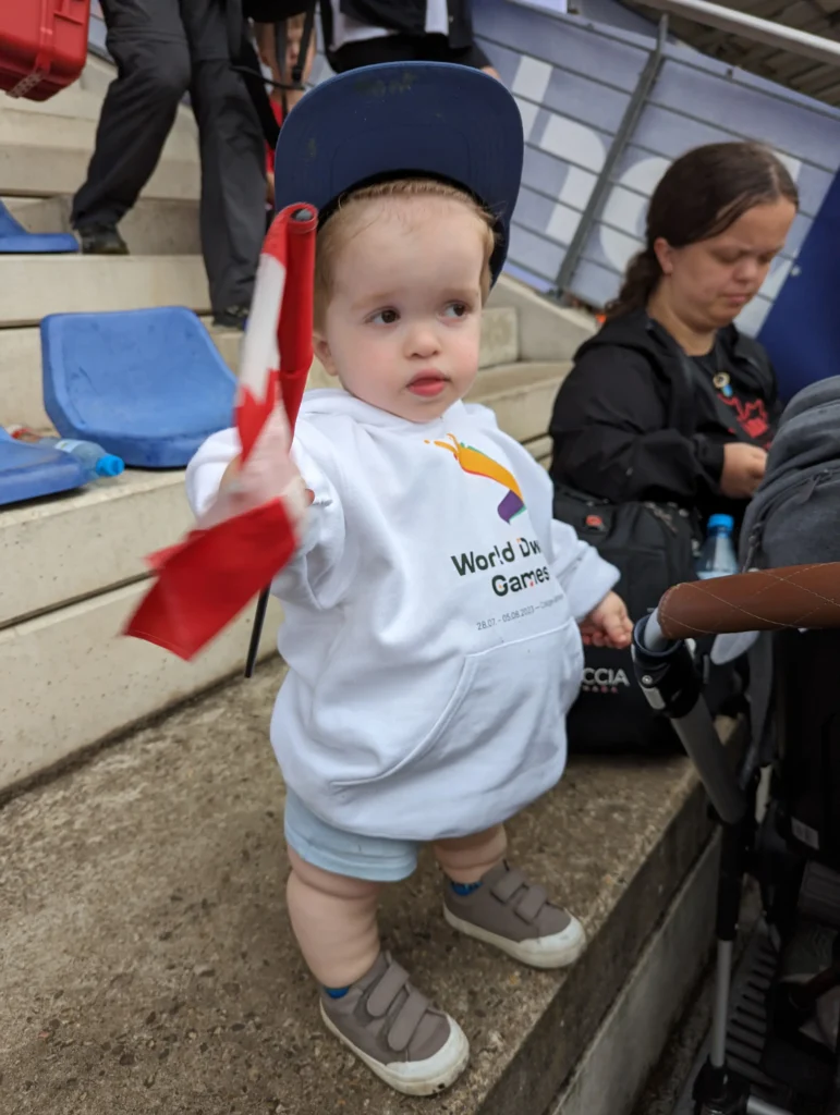A toddler in a white hoodie and blue cap stands on a stadium step, waving a red and white flag, surrounded by cheering fans.