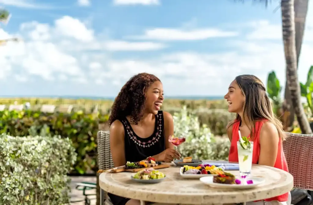 Two women enjoying tropical cocktails and a colorful lunch at a sunny outdoor beachside restaurant with palm trees and ocean views.