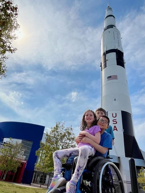 A child in a wheelchair sits in front of a towering rocket against a blue sky, with a modern building and trees nearby.