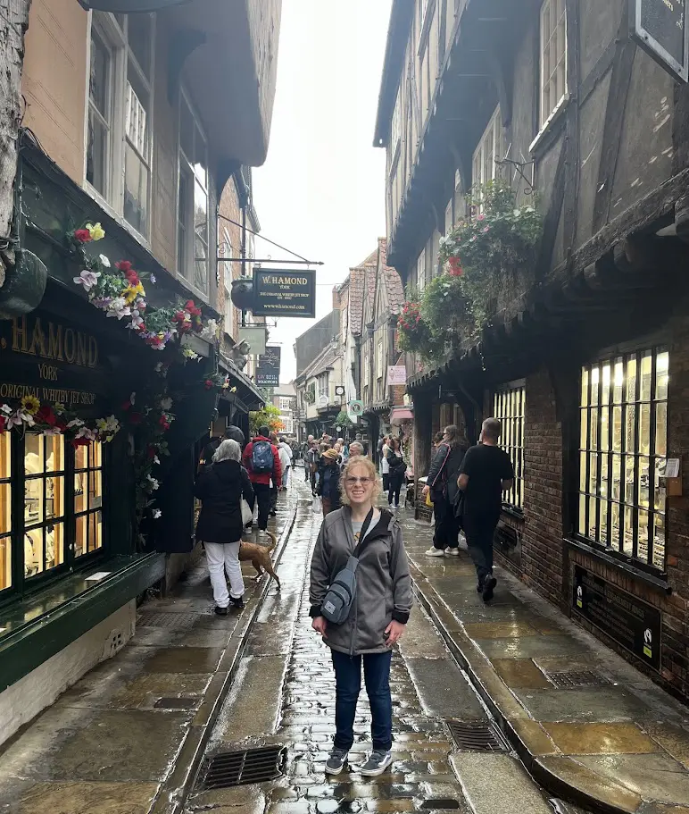 A smiling woman stands on a wet cobblestone street lined with historic timber-framed buildings and flower-decorated shopfronts in The Shambles, York, England, surrounded by other tourists on a cloudy day.