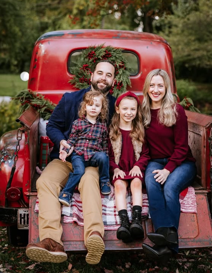A family of four sits together on the back of a vintage red truck, adorned with a festive wreath, surrounded by autumn foliage.