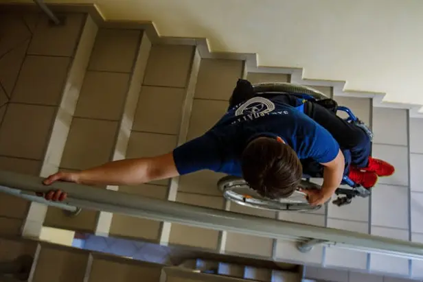 Overhead view of a person in a wheelchair navigating down a flight of stairs, holding the railing. The setting is challenging but conveys determination.