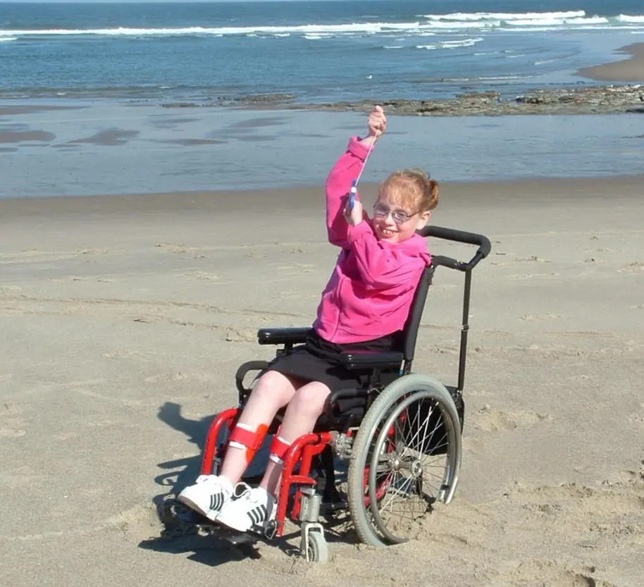 Young girl in a wheelchair smiling and flying a kite on a sandy beach near the ocean under a clear blue sky.