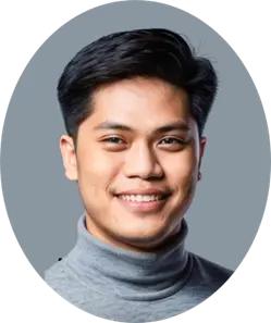 A person with short dark hair smiles warmly, wearing a gray turtleneck against a neutral gray background. The mood is friendly and approachable.