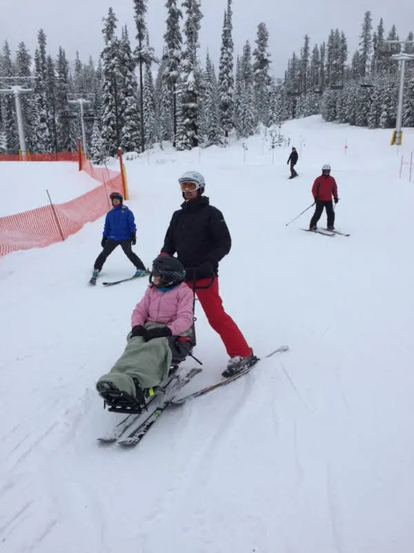 Person in a sit-ski being guided by an instructor on a snowy mountain slope with other skiers in the background.