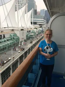 Smiling woman standing on a cruise ship balcony with a view of a modern port terminal and high-rise buildings in the background.