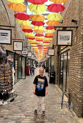 Woman standing in a vibrant shopping alley decorated with colorful hanging umbrellas, surrounded by boutique stores and cobblestone pavement.