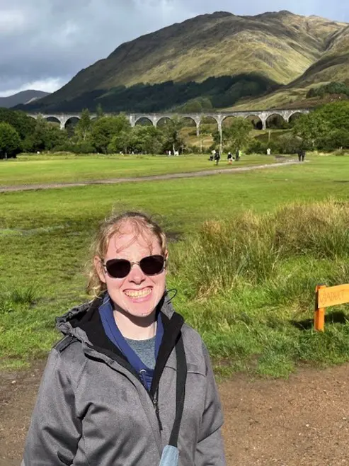 A smiling woman wearing sunglasses and a gray jacket stands in front of the scenic Glenfinnan Viaduct in Scotland, with lush green fields and a mountainous backdrop under a partly cloudy sky.