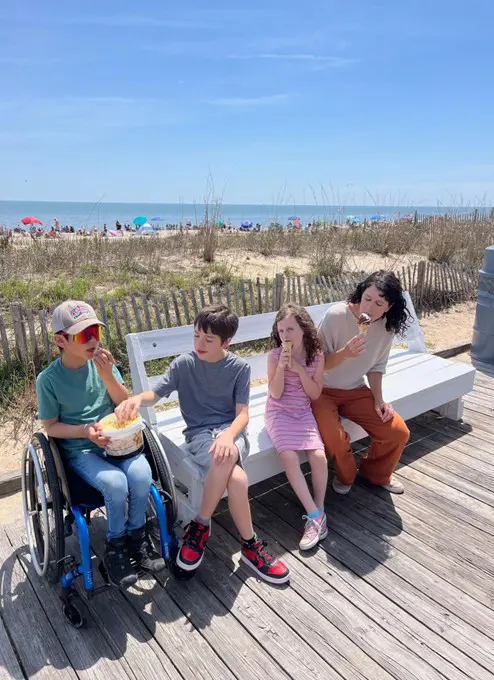 A group of children sits on a bench at the beach, enjoying snacks with sunny skies and ocean waves in the background.