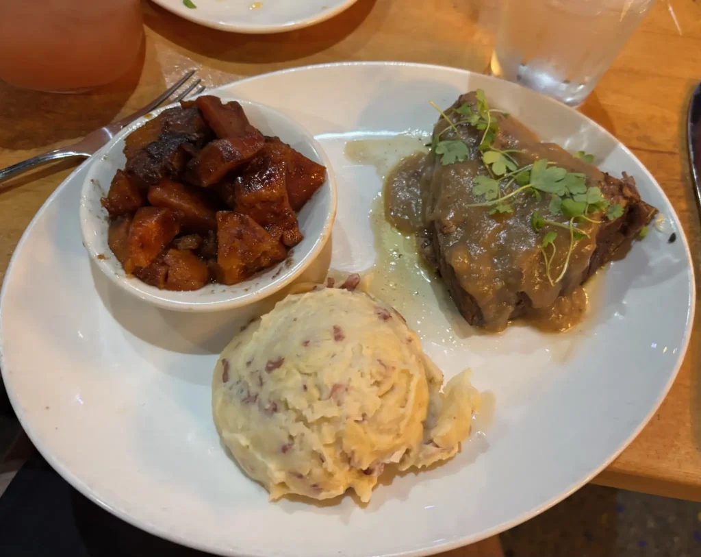 A hearty meal featuring glazed sweet potatoes, creamy mashed potatoes, and a beef dish with gravy and herbs on a white plate, exuding a comforting tone.