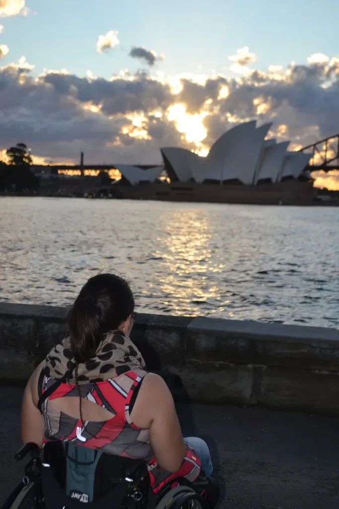 A person in a wheelchair gazes at the Sydney Opera House and sunset, reflecting on the water, surrounded by clouds.