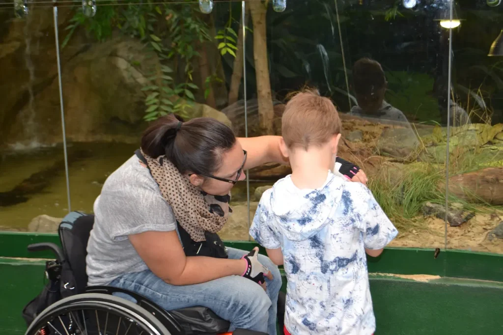 A person in a wheelchair interacts with a child at an aquarium exhibit, surrounded by lush greenery and a water feature.