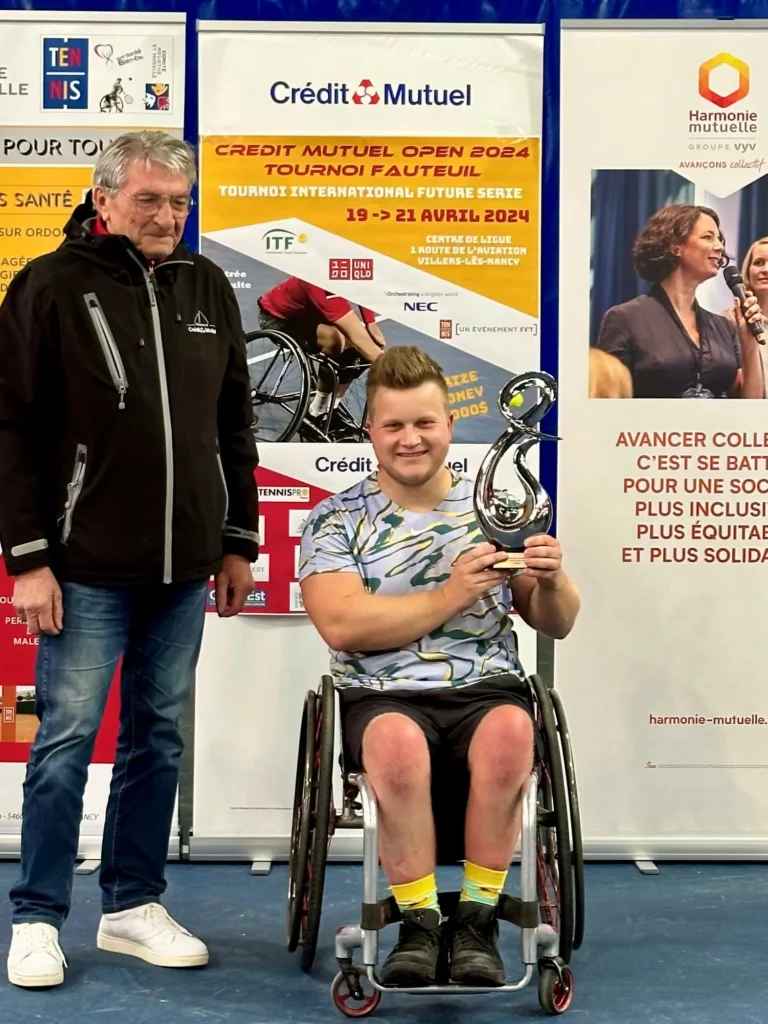 A smiling man in a wheelchair holds a trophy, posing next to an older man standing. They are in front of banners for a wheelchair tennis tournament.