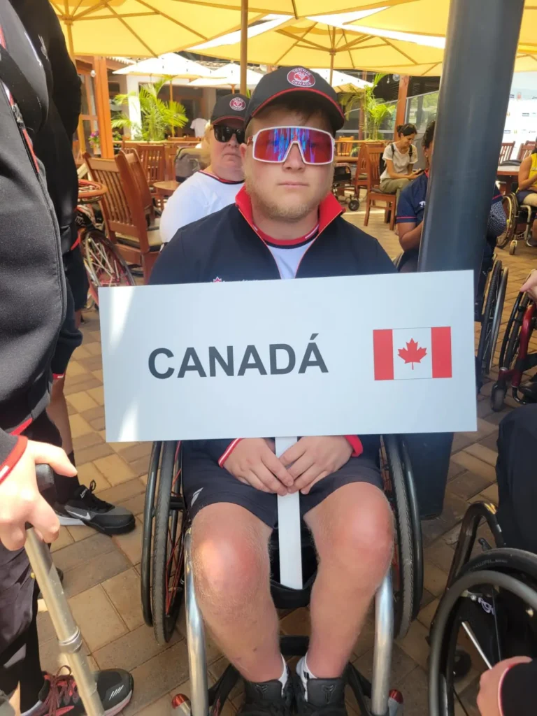 A man in a wheelchair holds a sign with the word Canadá and a Canadian flag. He wears sunglasses, a black cap, and a jacket, and is surrounded by others under large patio umbrellas.