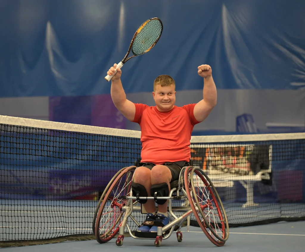 A male wheelchair tennis player in a gray tank top hits a tennis ball with a racket on an outdoor court. A black banner and chain-link fence are in the background.