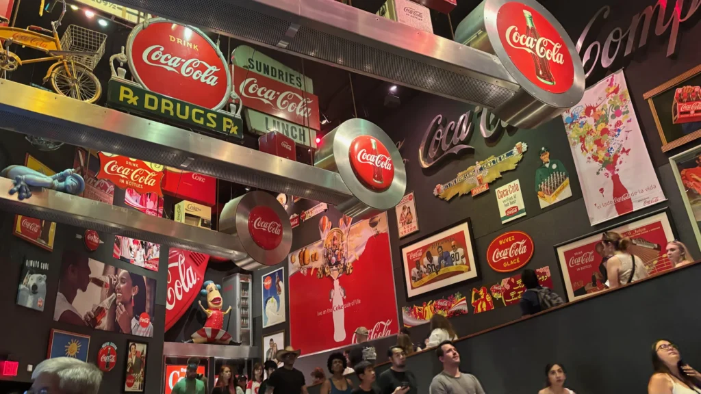 Wall filled with vintage Coca-Cola signs and memorabilia, including logos and colorful posters. People walking and looking around, creating a nostalgic atmosphere.