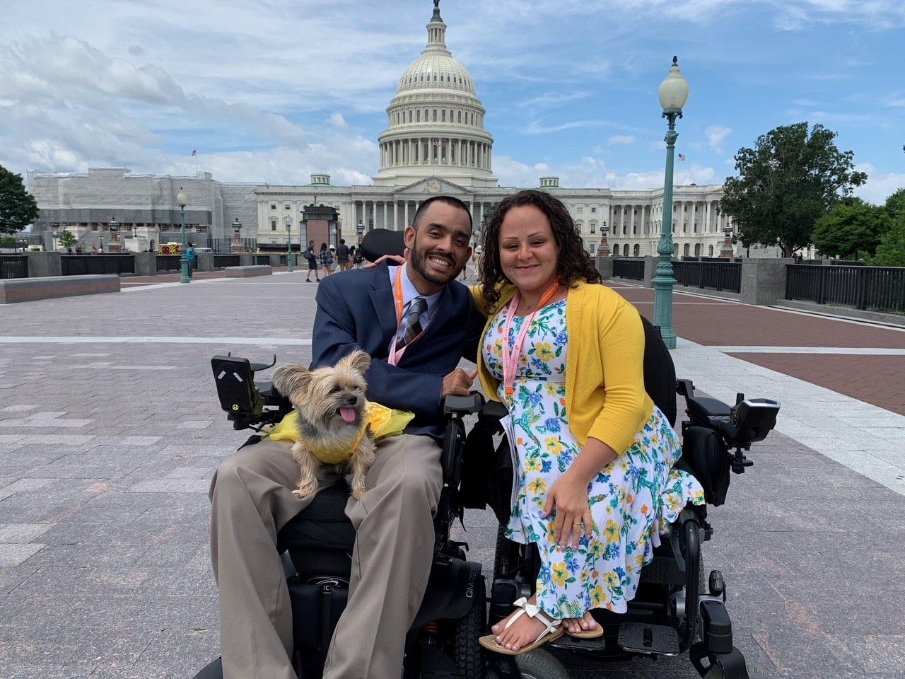 A couple in wheelchairs poses for a photo in front of the U.S. Capitol building in Washington, D.C. The man wears a dark business suit with an orange lanyard, and the woman wears a floral dress with a yellow cardigan. A small Yorkshire Terrier wearing a yellow vest sits on the man's lap. The iconic Capitol dome and building are clearly visible in the background under a partly cloudy sky.