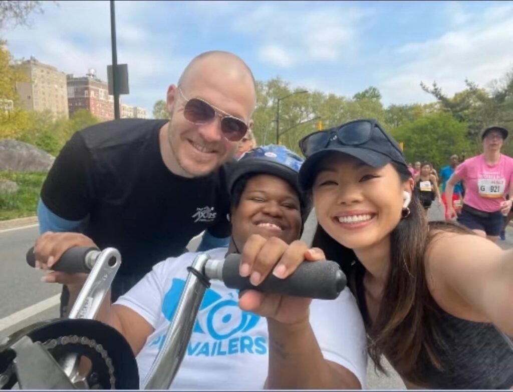 Three people pose together at what appears to be a running event or race. A bald man in sunglasses and a black athletic shirt stands behind two women - one Black woman in a white "VAILERS" t-shirt and cap, and one Asian woman in dark athletic wear with a cap. The setting appears to be in a park with trees and other participants visible in the background.
