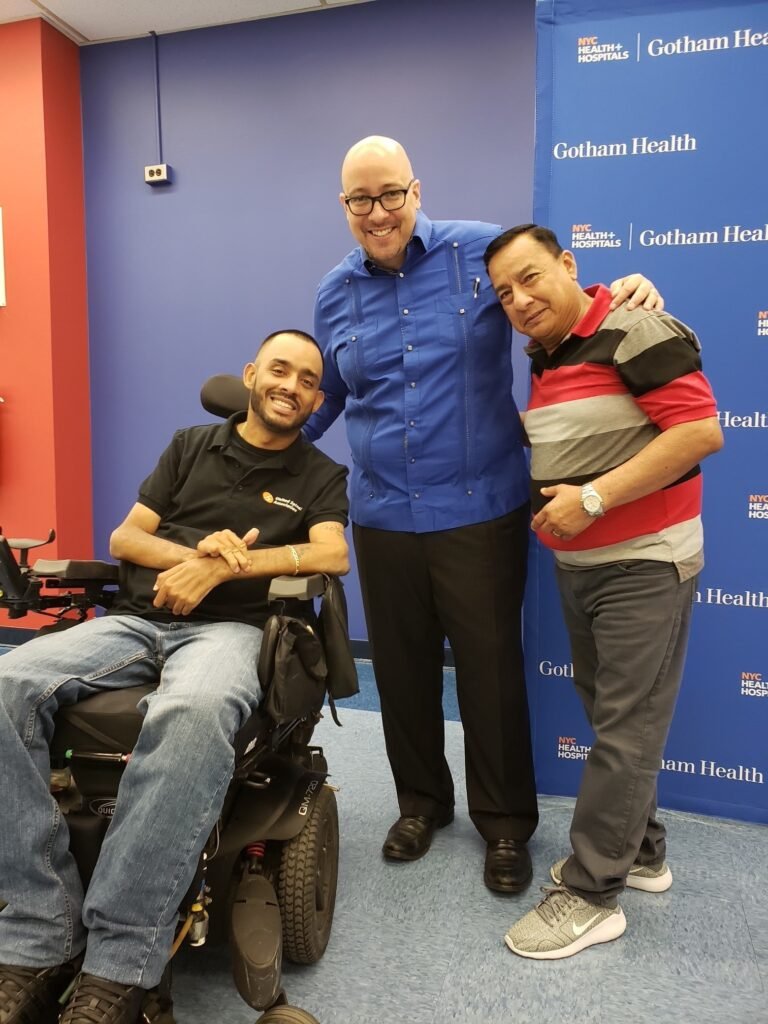 Three individuals are posing together in a healthcare setting, with a Gotham Health backdrop and wheelchair visible in the foreground.